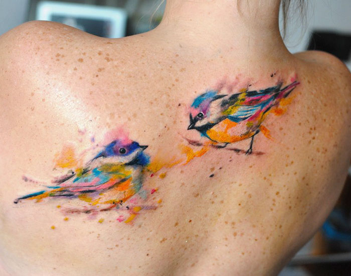 Colorful watercolored small bird tattoos on a person's upper back.
