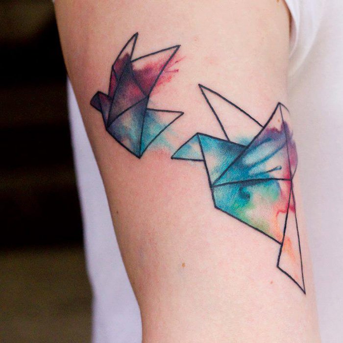 Colorful origami small bird tattoos on arm, featuring abstract watercolor design.