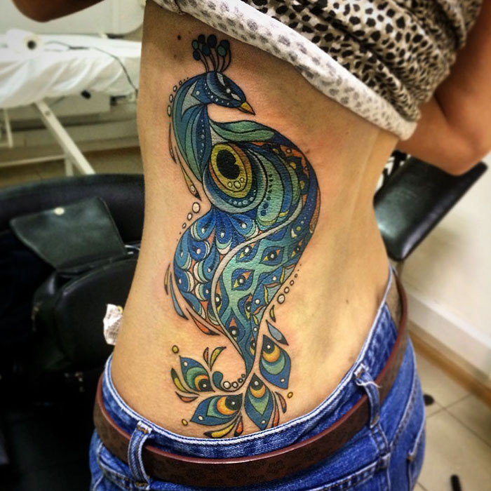 Colorful small bird tattoo on a person's side, featuring intricate peacock-inspired patterns.