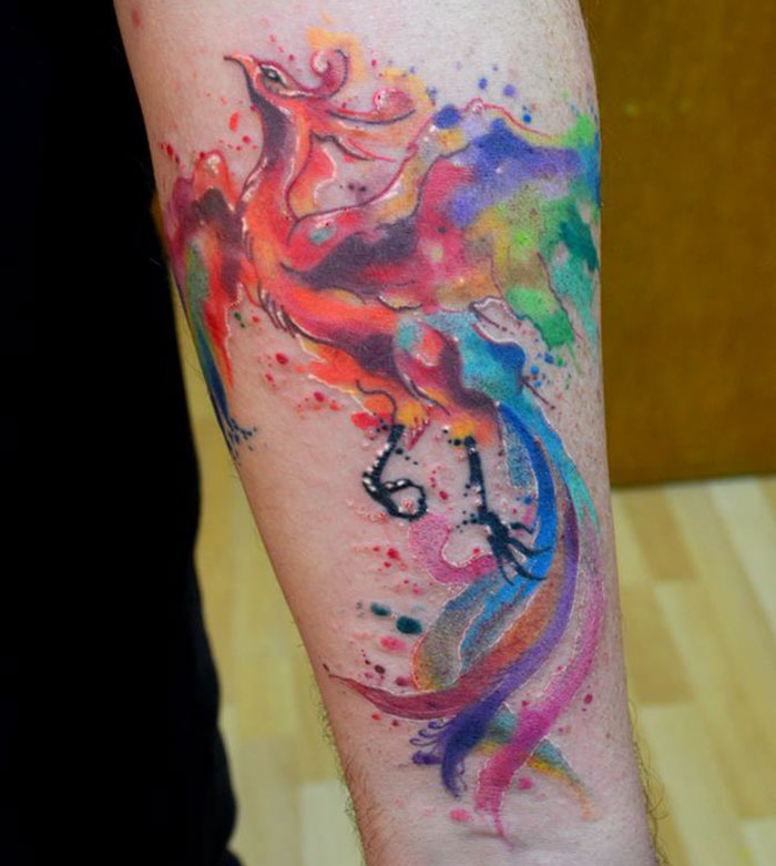 Vibrant watercolor small bird tattoo on forearm featuring colorful feathers and dynamic design.