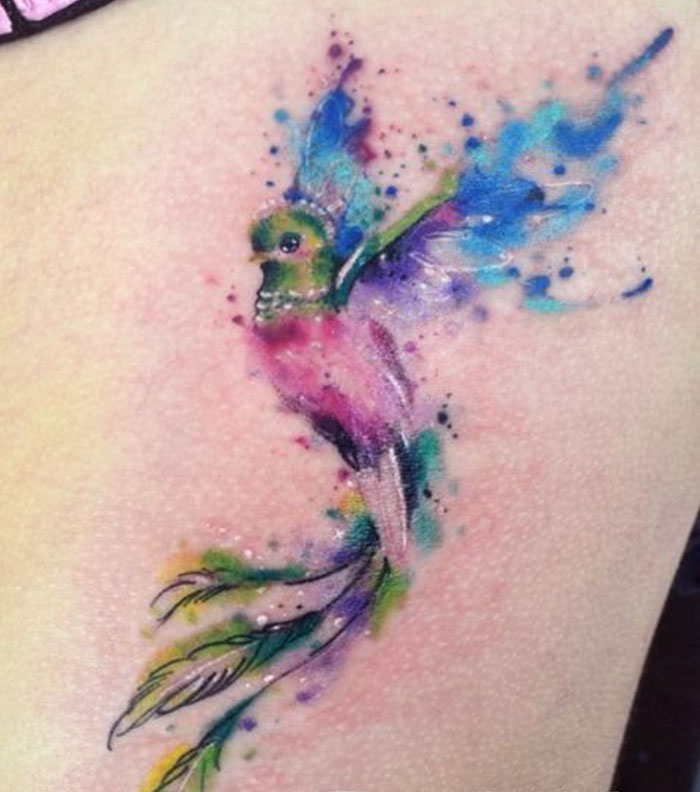 Colorful watercolor small bird tattoo with vibrant blue and pink feathers.