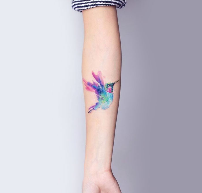 Colorful small bird tattoo on forearm, vibrant watercolor design in purple, blue, and green tones.