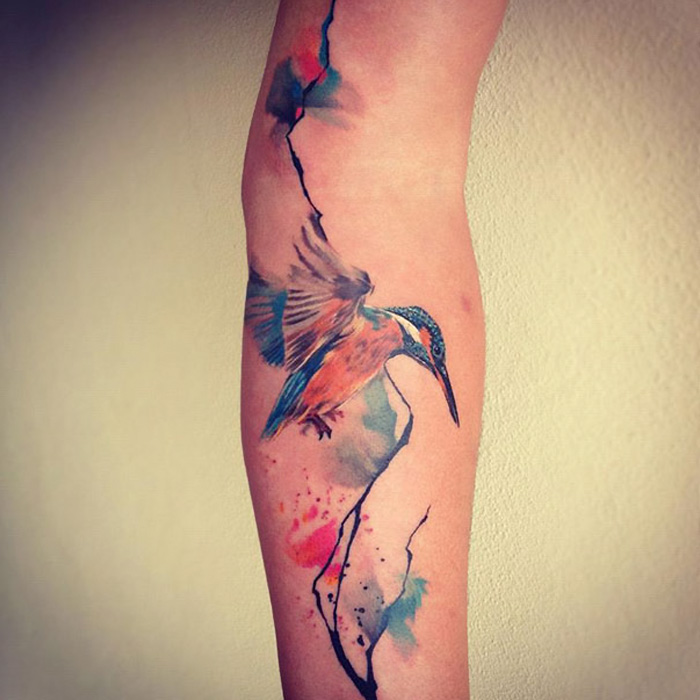 Watercolor small bird tattoo with colorful ink splashes on arm.