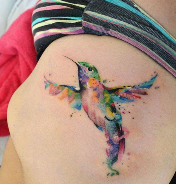 Colorful small bird tattoo on shoulder, featuring a vibrant design with splashes of watercolor style.