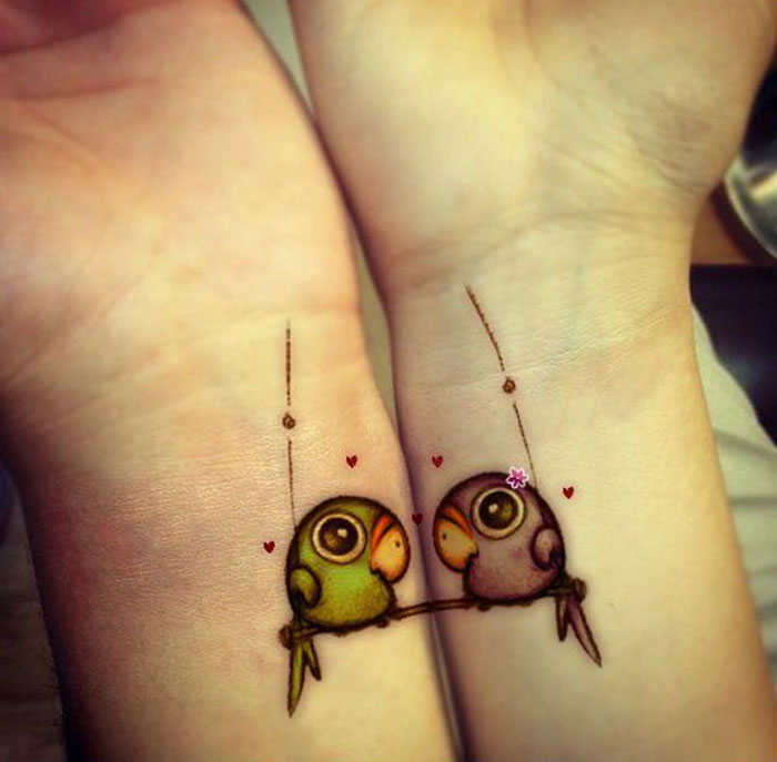 Two colorful small bird tattoos on wrists, featuring heart accents and intricate details.