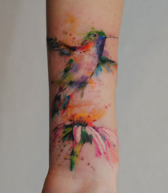Colorful watercolor small bird tattoo on forearm, capturing vibrant artistry and inspiration.