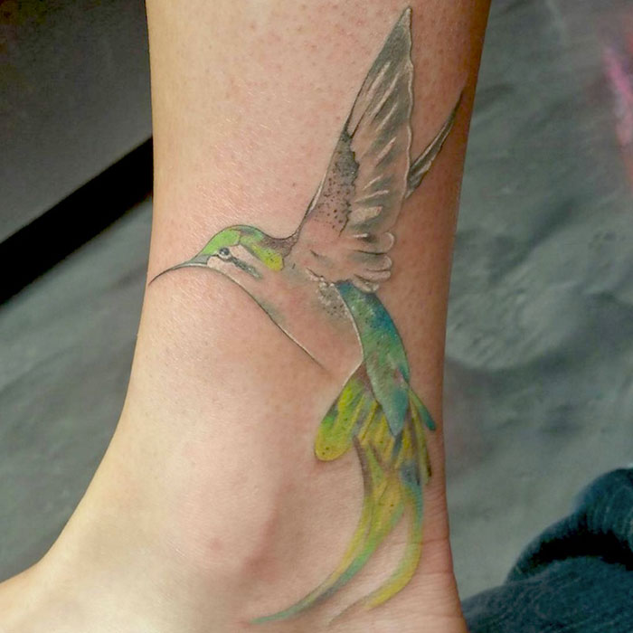 Colorful small bird tattoo on ankle, showcasing intricate wing and feather designs with vibrant hues.
