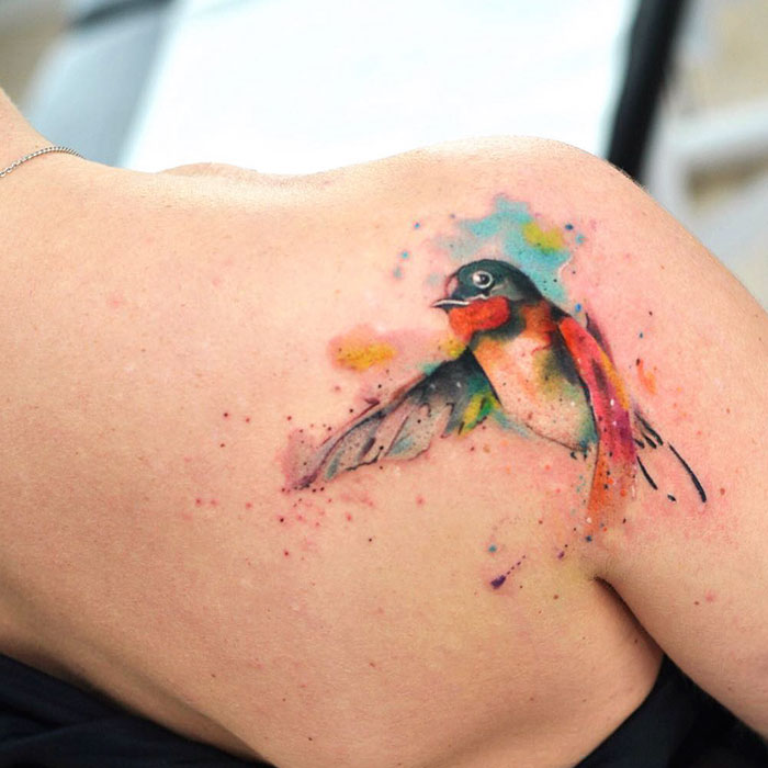 Colorful small bird tattoo on shoulder, showcasing a vibrant design with watercolor effects.