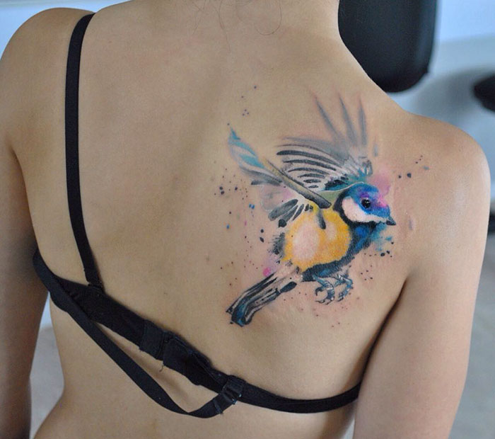 Colorful small bird tattoo on a woman's back, showcasing unique watercolor design.