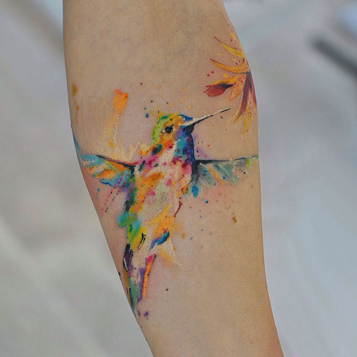 Colorful watercolor small bird tattoo on forearm with vibrant hues.
