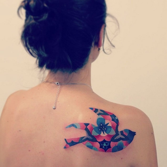 Colorful small bird tattoo design on woman's back, featuring intricate floral patterns.