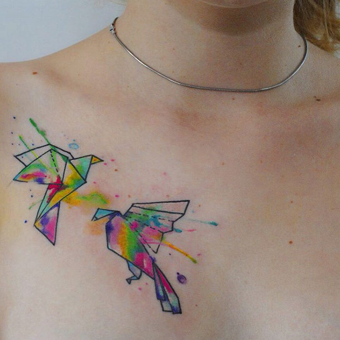 Colorful origami-style small bird tattoo on a person's chest, featuring vibrant splashes of ink.