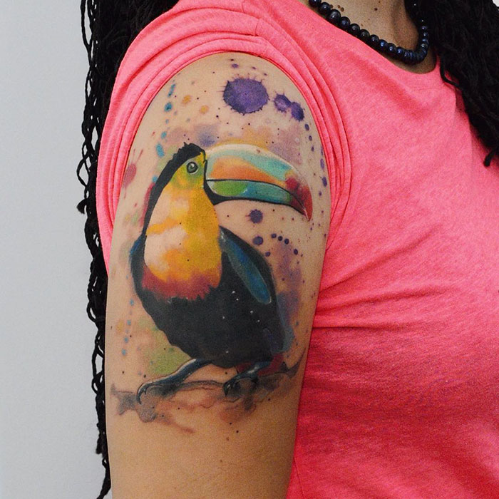 Colorful small bird tattoo featuring a toucan on a person's arm in a pink shirt, inspired ink art masterpiece.