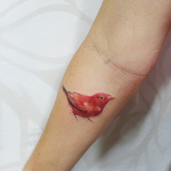 Small bird tattoo on forearm, featuring a vibrant red bird with a piercing yellow eye.
