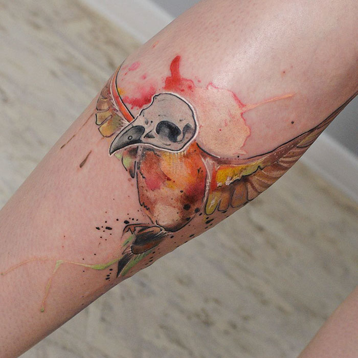 Unique small bird tattoo with artistic watercolor style on leg, featuring a vibrant design and a skull motif.