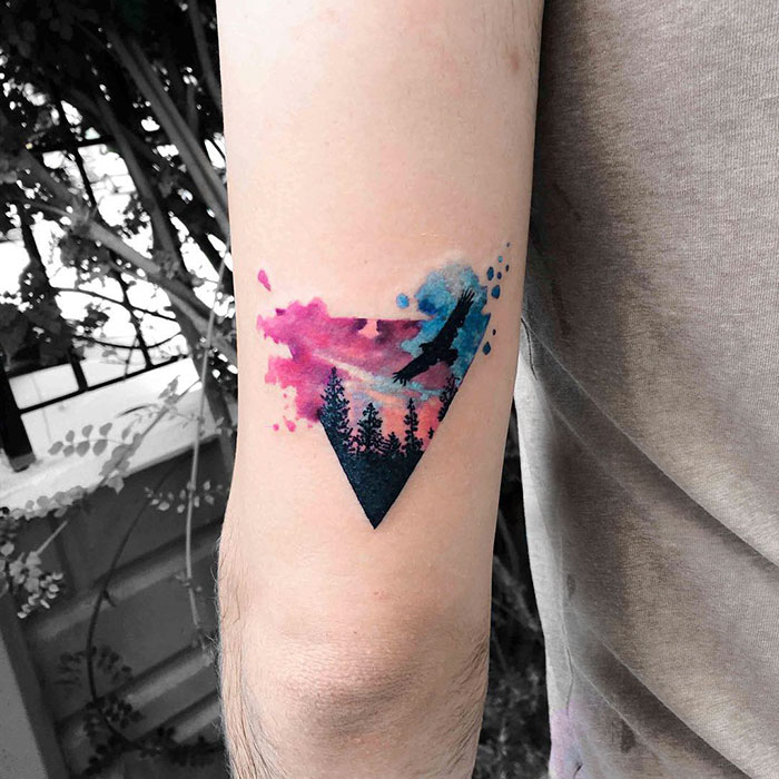 Colorful small bird tattoo with forest and sky design, featuring vibrant hues on arm.