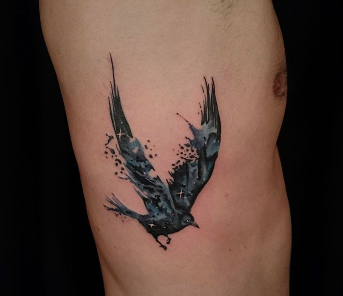 Watercolor style small bird tattoo on side, featuring a flying blue-black bird with dynamic brush strokes.