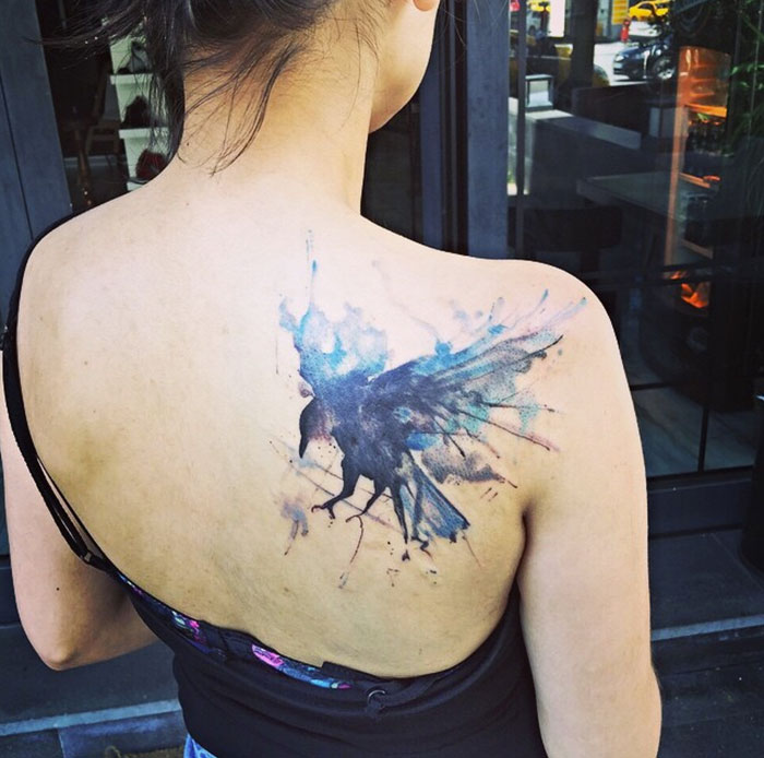 A woman's shoulder with a watercolor-style small bird tattoo, featuring vibrant blue and black splashes.