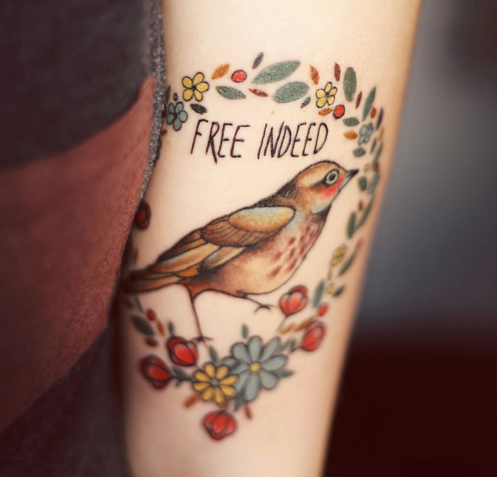 Small bird tattoo with the words "Free Indeed" surrounded by flowers, showcasing unique bird tattoo art.