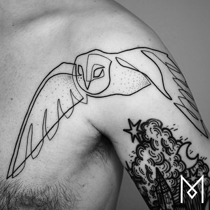 Unique small bird tattoo in black ink on a person's shoulder, featuring an abstract design of an owl in flight.
