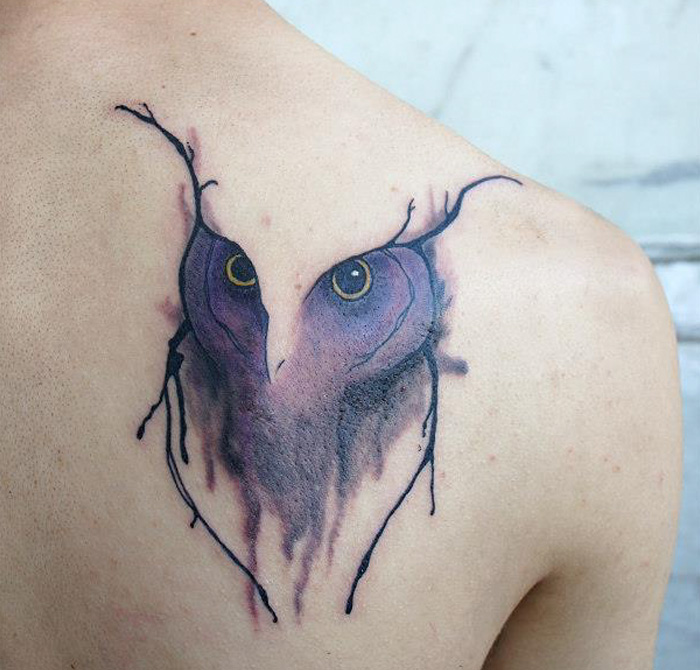 Unique small bird tattoo design of an owl face on shoulder with abstract brushstroke effect.
