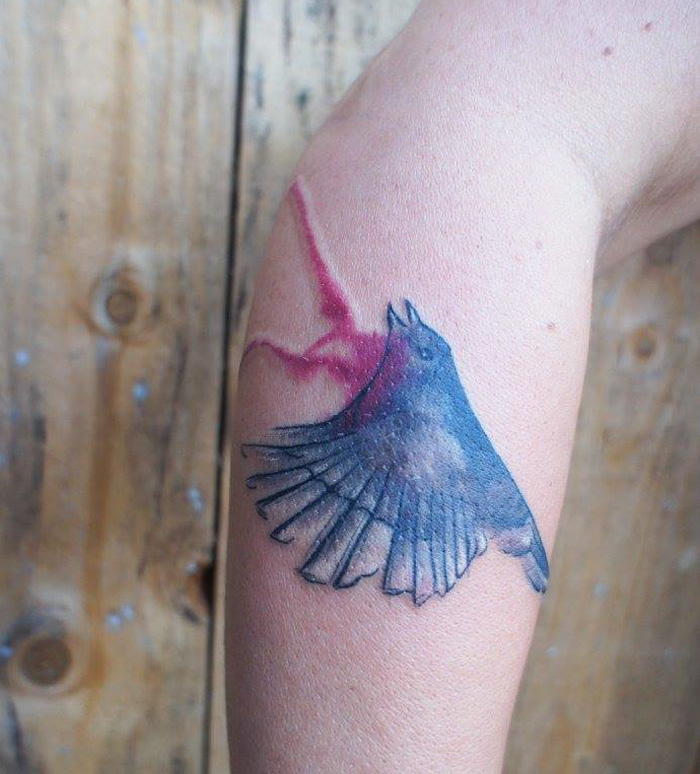 Colorful small bird tattoo on forearm with abstract background.