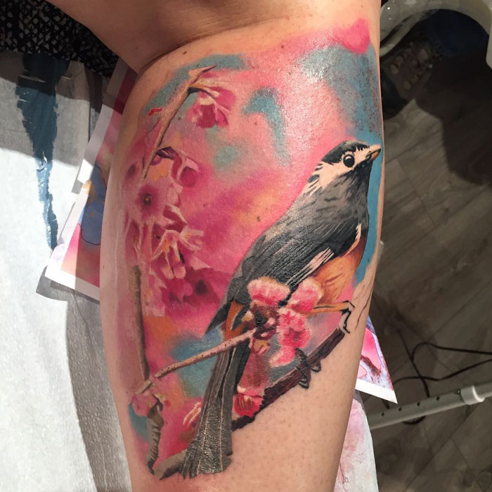 Colorful tattoo of a small bird perched on pink blossoms, with a vibrant watercolor background.