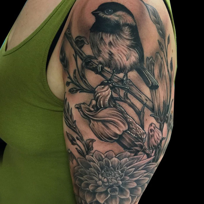 Arm tattoo featuring a detailed small bird perched on flowers and branches in black ink.