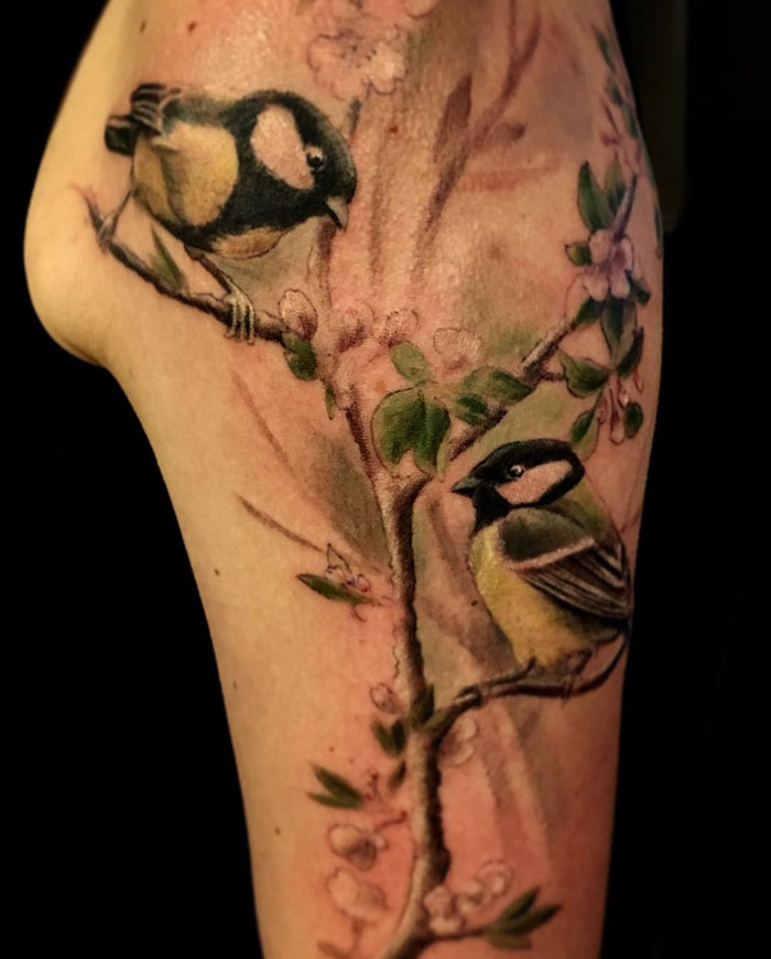 Small bird tattoo depicting two birds on a blossoming branch, showcasing intricate and vibrant details.