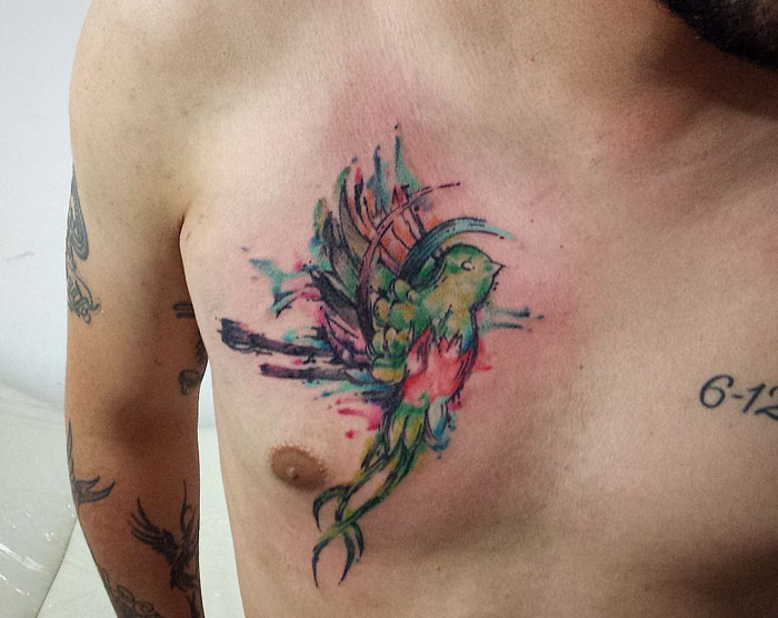 Colorful small bird tattoo on a man's chest, featuring vibrant watercolor style.