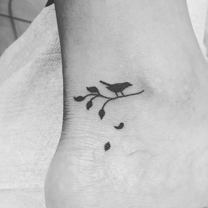 Small bird tattoo on ankle with minimalist branch design.