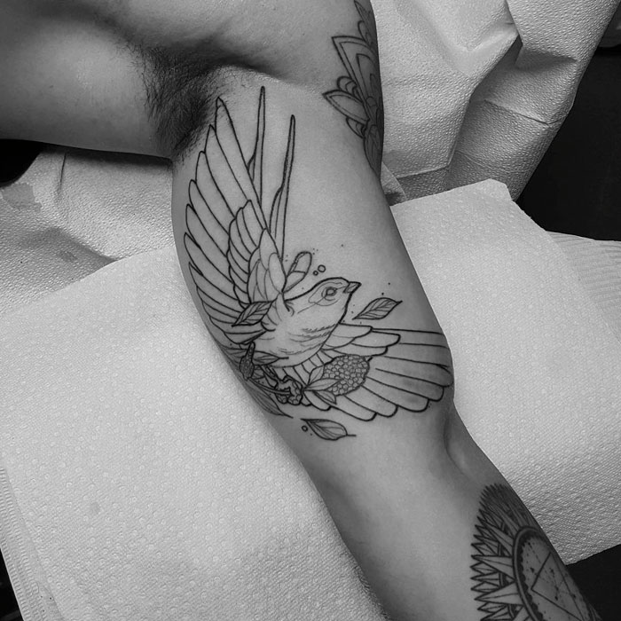 Black and white small bird tattoo design on an arm, showcasing intricate linework and floral details.