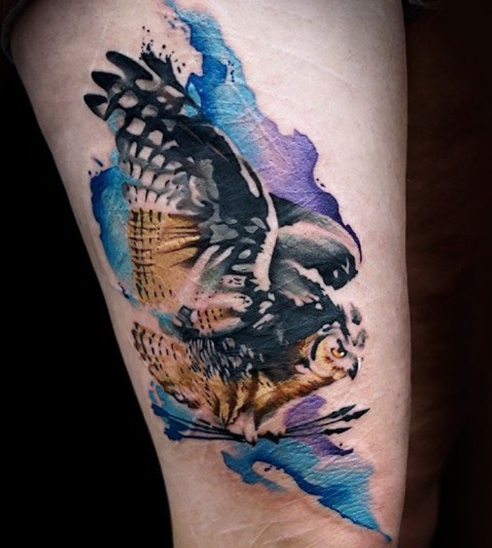 Watercolor style small bird tattoo with vibrant blue and purple accents on a person's arm.