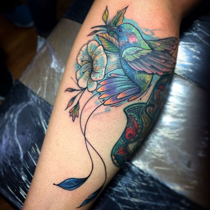 Colorful small bird tattoo with floral elements on a person's leg.