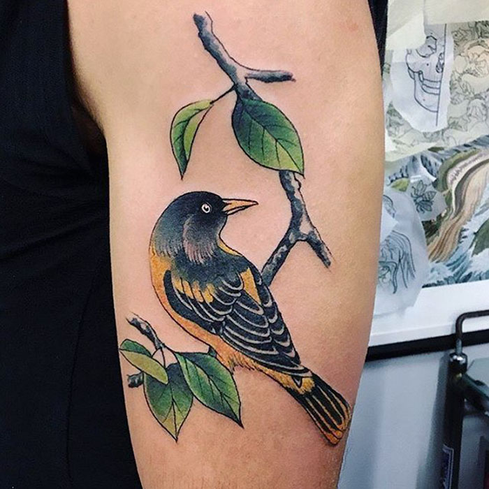 Colorful small bird tattoo on an arm, featuring intricate details with a branch and leaves.