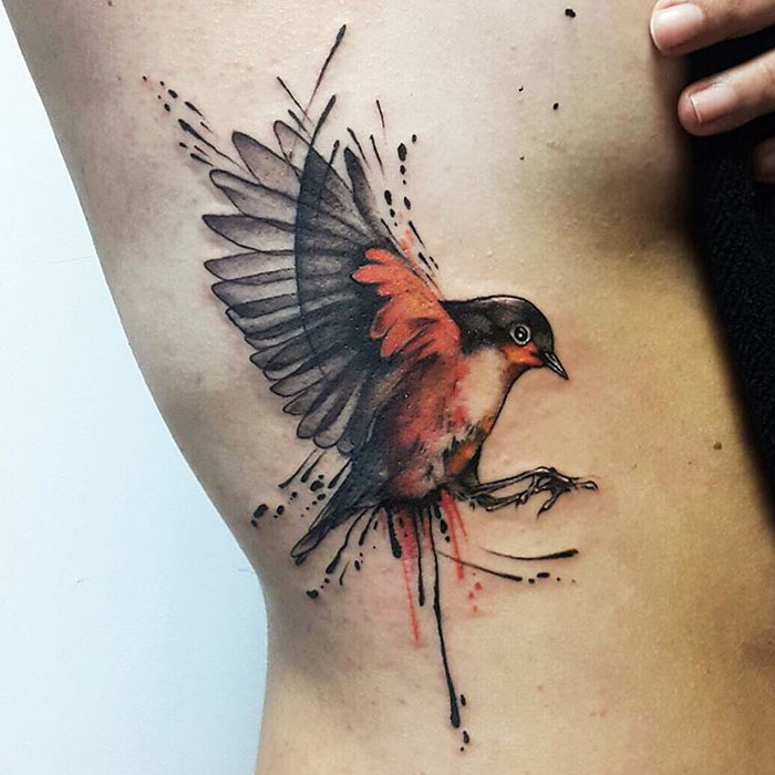 Unique small bird tattoo with vivid colors and artistic splatter design on the side of the body.