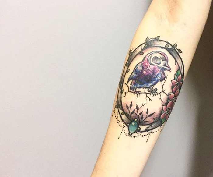Colorful small bird tattoo on forearm, featuring floral and gemstone details.