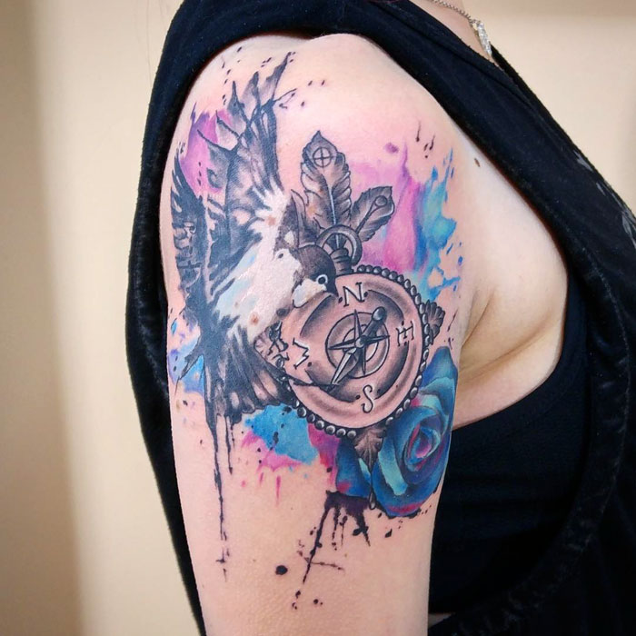 Unique small bird tattoo with a compass, feathers, and watercolor accents on arm.