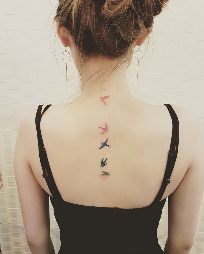 Woman with small bird tattoos in various colors on her back, wearing a black top, showcasing unique bird tattoo designs.