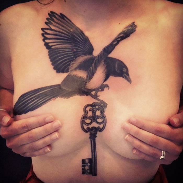 Small bird tattoo design featuring a magpie with a key, inked on a person's chest.