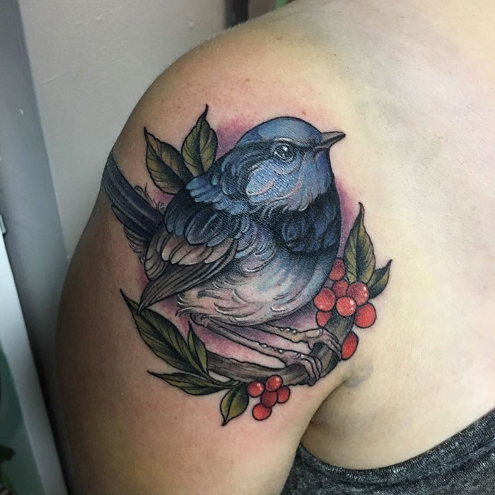 Blue small bird tattoo on shoulder with leaves and red berries, showcasing unique small bird tattoo artistry.