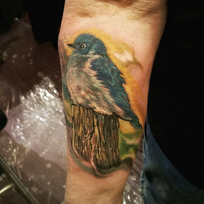 Colorful small bird tattoo on forearm, featuring a blue bird perched on a wooden stump, showcasing intricate details.