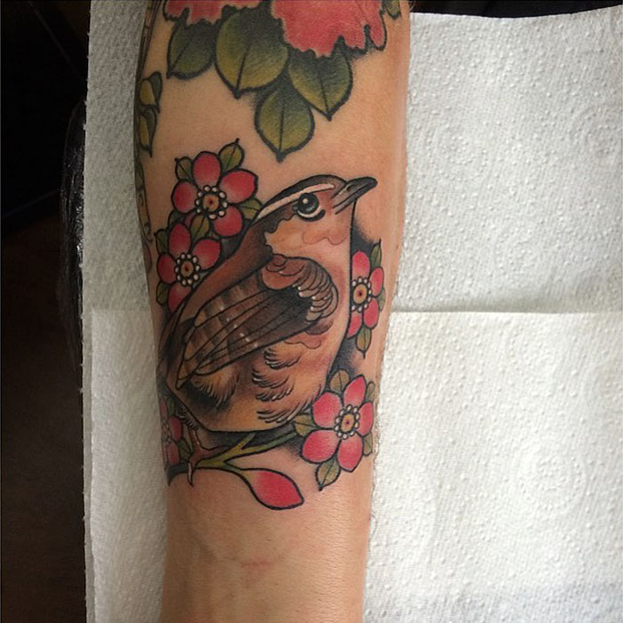 Colorful small bird tattoo among flowers on forearm, showcasing unique tattoo artistry.