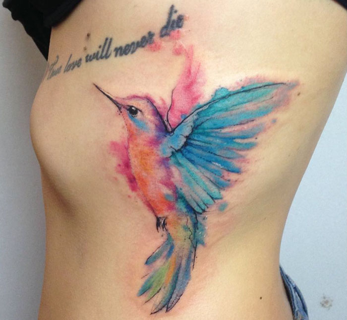Colorful small bird tattoo with watercolor effect on side of the torso.