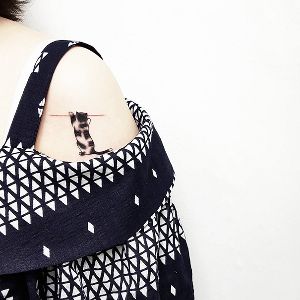 Cat tattoo on shoulder featuring a black cat hanging from a red line, under patterned black and white garment.