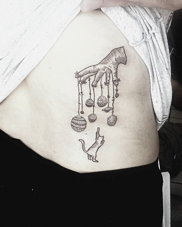 Cat tattoo design with a playful cat reaching towards dangling balls of yarn on someone's side.