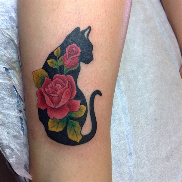 Cat tattoo design featuring a black silhouette of a cat filled with vibrant red roses and green leaves.