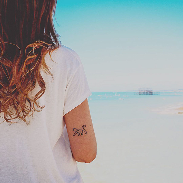 A woman with a minimalist cat tattoo on her arm, standing by the beach.