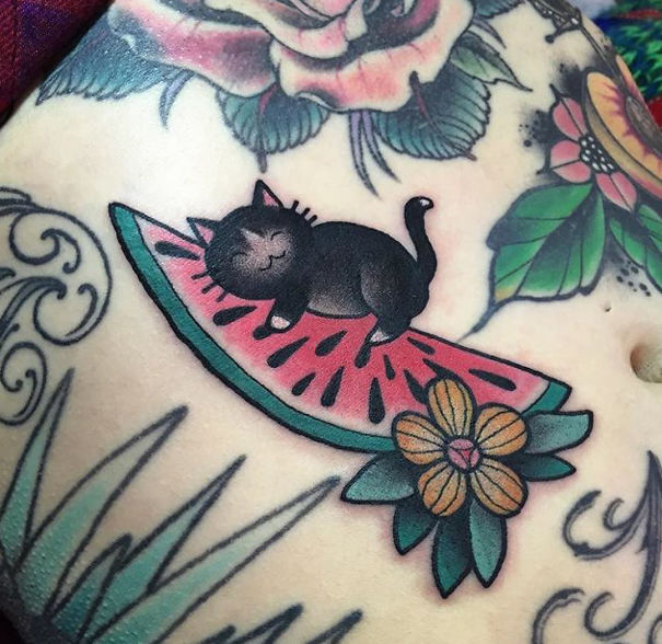 Cat tattoo of a black cat lying on a watermelon slice, surrounded by colorful flowers.