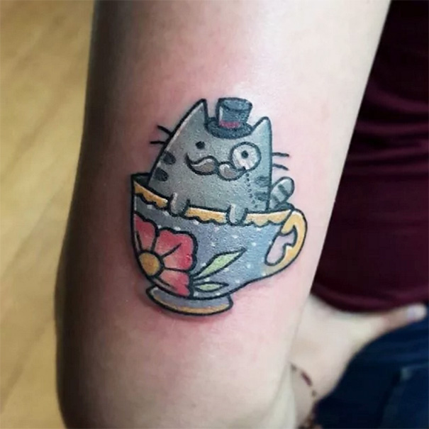 Cat tattoo featuring a cartoon cat with a monocle and top hat inside a floral teacup.